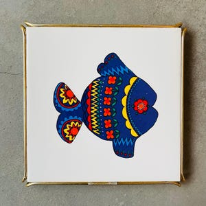 Może przedstawiać: Niebieska i żółta ryba z czerwonymi i żółtymi kwiatami na białym tle. Ryba jest oprawiona w złoto.