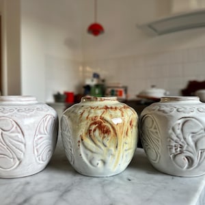 Könnte beinhalten: Drei Keramikvasen mit runder Form und geprägten floralen Designs. Die Vasen sind in Weiß-, Creme- und Brauntönen gehalten. Die Vasen werden auf einer Marmorarbeitsplatte ausgestellt.