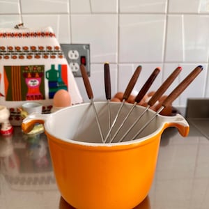 Puede incluir: Una olla para fondue de esmalte color naranja brillante con dos asas laterales y un interior blanco. Cinco tenedores de metal para fondue con mangos de madera marrón oscuro están colocados dentro. Cada mango tiene una pequeña punta de color amarillo, verde, naranja, rojo o azul.