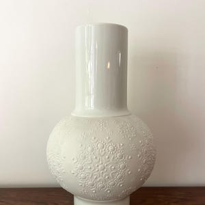 May include: A white ceramic vase with a round, embossed body and a cylindrical neck. The vase features a delicate snowflake pattern.