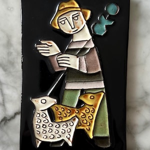 Può includere: Piastrella in ceramica rettangolare con sfondo nero raffigurante un'illustrazione stilizzata. Una figura con cappello e camicia a righe è in piedi con una pecora e un pesce. La pecora è bianca con macchie nere e il pesce è giallo.