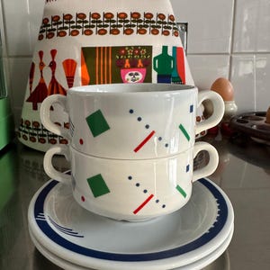 Op de afbeelding: Twee witte soepkommen met handvatten, versierd met rode, groene en blauwe geometrische vormen, gestapeld op schotels met een blauwe rand. Een theedoek met patroon staat op de achtergrond.