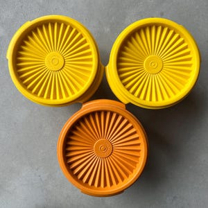 Könnte beinhalten: Draufsicht auf drei Kunststoffdeckel für Lebensmittelbehälter. Zwei sind leuchtend gelb und einer orange. Jeder Deckel ist rund mit einem Sonnenstrahl-Design und einer kleinen kreisförmigen Vertiefung in der Mitte. Die Deckel liegen auf einer grauen Oberfläche.