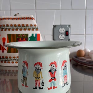 May include: A vintage, light blue enamelware pot with a decorative rim. The pot features four cartoon gnomes in colorful outfits. Each gnome is holding a lantern. The pot is likely used for cooking or storage.