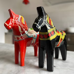 Pode incluir: Dois cavalos de Dala de madeira, artesanato tradicional sueco. Um é vermelho brilhante com detalhes pintados em branco, azul e amarelo. O outro é preto com padrões pintados coloridos semelhantes. Ambos estão em uma superfície branca.