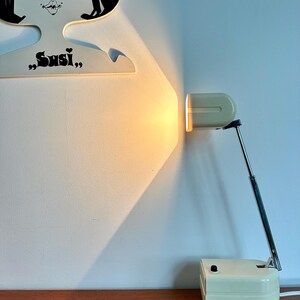 Könnte beinhalten: Eine cremefarbene Schreibtischlampe mit rechteckigem Kopf und verstellbarem Arm in Chromoptik, ausgestattet mit einem quadratischen Sockel und schwarzem Schalter. Die Lampe strahlt ein warmes gelbes Licht auf ein weißes Wandschild mit der Aufschrift Susi.
