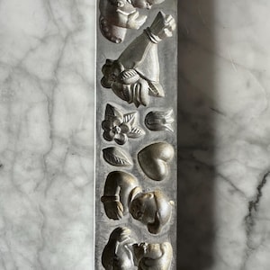 May include: A vintage, rectangular metal mold with detailed relief designs. The mold features images of a baby, flowers, leaves, and a heart. The text "V. LAERE LEIDEN HOLLAND No.1002" is embossed at the bottom. The mold has a silver-gray color.