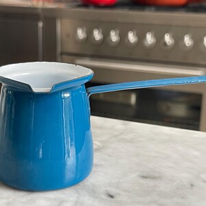 Könnte beinhalten: Eine blaue Emaille-Kaffeekanne mit weißem Innenraum und langem Griff. Die Kanne hat ein klassisches Design, geeignet für den Gebrauch auf dem Herd. Die Kanne steht auf einer Marmoroberfläche.