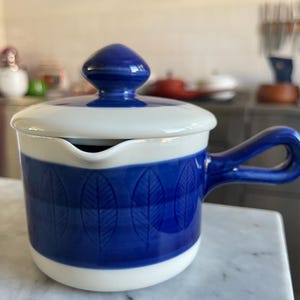 Pode incluir: Um cremoso de cerâmica branco e azul com tampa e alça. O cremoso tem um design de folhas azuis e brancas.