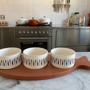 Może przedstawiać: Trzy białe ceramiczne miski z wzorem niebieskich i czerwonych ryb są ułożone na drewnianej, rybiej tacy. Taca znajduje się na białym marmurowym blacie.