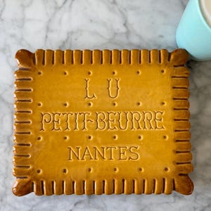 May include: A ceramic coaster in the shape of a biscuit, with the words "LU PETIT-BEURRE NANTES" embossed on the surface. The coaster is a golden-brown color and has a detailed edge design. A light blue mug with a white foamy drink is in the background.