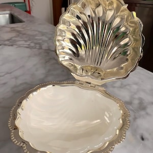 May include: A silver-toned metal clam shell dish with a hinged lid. The interior of the bottom shell is smooth and white, while the interior of the top lid features a reflective, scalloped, and ribbed design with a beaded edge along the rim.