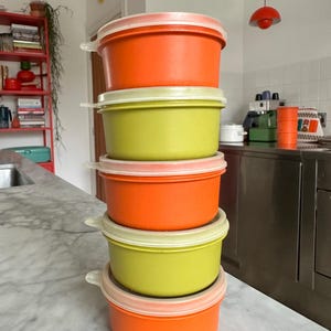 Op de afbeelding: Een stapel van vijf kleurrijke plastic voedselcontainers. De containers wisselen af tussen oranje en limoengroen, elk met een wit deksel. De containers staan op een witte marmeren aanrecht.