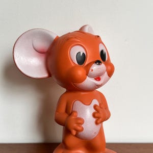 May include: A vintage orange Jerry Mouse figurine from the Tom and Jerry cartoon. The figure has large pink-lined ears, a white chest, and a small red tongue. The figurine is standing and has a smiling expression, reminiscent of the cartoon.