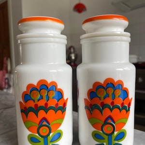 以下が含まれることがあります： オレンジ色の蓋が付いた白いガラス瓶のペア。各瓶には、オレンジ、青、緑の大きなカラフルな花の模様があります。この瓶はレトロでヴィンテージな美しさがあり、キッチンの収納やディスプレイに最適です。
