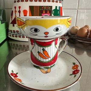 以下が含まれることがあります： 気まぐれなセラミックティーカップとソーサーのセット。ティーカップは、黄色、オレンジ、白のディテールで猫の顔としてデザインされています。ソーサーは茶色の縁と花のアクセントがあります。ティーカップは、お揃いのソーサーの上に置かれています。