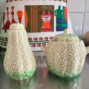 May include: A vintage ceramic salt shaker and sugar bowl set. The salt shaker is pear-shaped, and the sugar bowl is round with a lid. Both pieces have a light beige, textured surface with a green base. The set is likely from the mid-century.