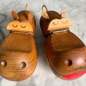 May include: Two wooden hippo-shaped boxes with hinged lids. The boxes are made of light brown wood with darker wood grain. The hippos have small ears, and the lids are painted with eyes and a mouth. One hippo has closed eyes, and the other has eyelashes.