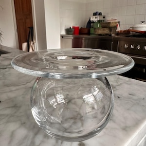 Puede incluir: Un florero de vidrio transparente con una base esférica y un borde circular ancho y plano en la parte superior. El recipiente está construido de vidrio transparente con una superficie lisa y reflectante, mostrando un contraste distintivo entre el cuerpo redondeado y la abertura horizontal.