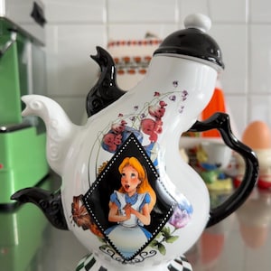 Pode incluir: Uma chaleira de cerâmica branca com detalhes em preto e um design extravagante. A chaleira apresenta uma imagem em forma de diamante de Alice no País das Maravilhas, padrões florais e uma base quadriculada.