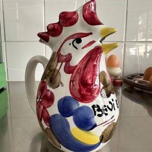 以下が含まれることがあります： 白いセラミックピッチャー、鶏のデザイン。 ピッチャーは赤い鶏冠、黄色のくちばし、青と赤の花柄が特徴です。 側面には「Bevi」の文字が書かれています。 ピッチャーには湾曲したハンドルが付いています。
