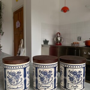 May include: Three white ceramic spice jars with blue and white floral designs and wooden lids. The jars are labelled "Nootmuskaat", "Kruidnagel", and "Foelie".