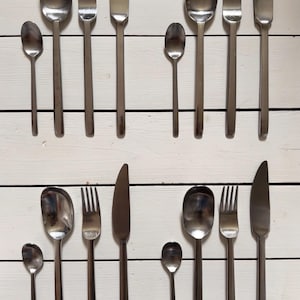 Pode incluir: Um conjunto de dezasseis peças de talheres em aço inoxidável dispostas em quatro grupos idênticos. Cada grupo consiste numa colher pequena, uma colher grande, um garfo e uma faca de mesa, todos com acabamento prateado polido e liso.