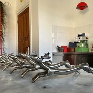 Puede incluir: Un juego de seis servilleteros plateados con forma de reno. Los renos están en posición de carrera, con cornamentas y patas detalladas. Están dispuestos en fila sobre una encimera de mármol, adecuados para mesas festivas.