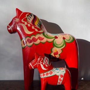 May include: Two traditional Swedish Dala horses, one large and one small, are painted in bright red with intricate white, yellow, green, and blue designs. The horses are standing on a wooden surface.