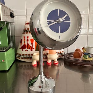 Op de afbeelding: Een bolvormige bureauklok van chroom met een flexibele metalen hals en een ronde voet. De wijzerplaat is wit met een blauwe cirkel in het midden, zilveren wijzers en een kleine letter t onderaan. De klok is gemonteerd op een dunne, spiraalvormige stang.