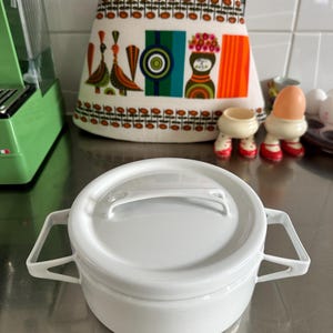 Puede incluir: Una olla de esmalte blanco con tapa y dos asas. La olla es redonda y se encuentra sobre una superficie de acero inoxidable. En el fondo, un paño de cocina colorido con un diseño retro y un electrodoméstico verde.
