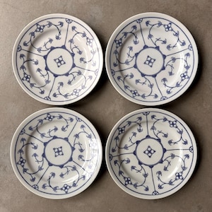 May include: Four white ceramic plates arranged in a square formation. Each plate features a central circular motif with a four-petaled design, surrounded by radiating blue lines and delicate, symmetrical floral patterns in a deep blue color.
