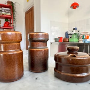 May include: Three brown ceramic kitchen canisters with lids. The canisters are cylindrical and have a textured, speckled finish. The set includes a tall, a medium, and a shorter, round container with a lid.