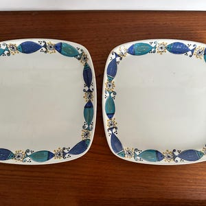 Può includere: Due piatti quadrati bianchi con un bordo di pesci blu e turchese e dettagli dorati. I piatti hanno angoli arrotondati e un design semplice, perfetti per servire antipasti o dessert.