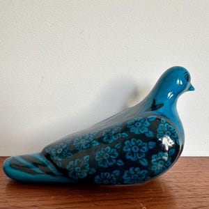 Op de afbeelding: Een keramieken duifbeeldje in tinten van turquoise en zwart. De vogel heeft een glanzende afwerking en is versierd met een bloemmotief op zijn vleugels en lichaam. Het beeldje staat op een houten oppervlak.