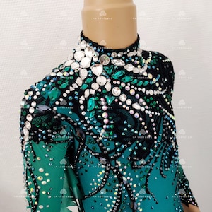 May include: A teal and black leotard with a high neckline, adorned with clear and emerald-green rhinestones. The garment features intricate lace detailing, suitable for performance or dance.