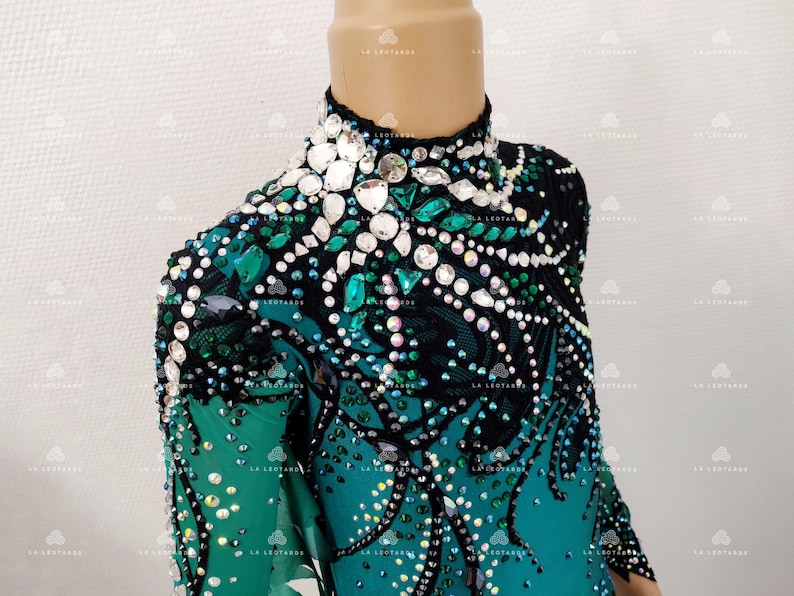 May include: A teal and black leotard with a high neckline, adorned with clear and emerald-green rhinestones. The garment features intricate lace detailing, suitable for performance or dance.