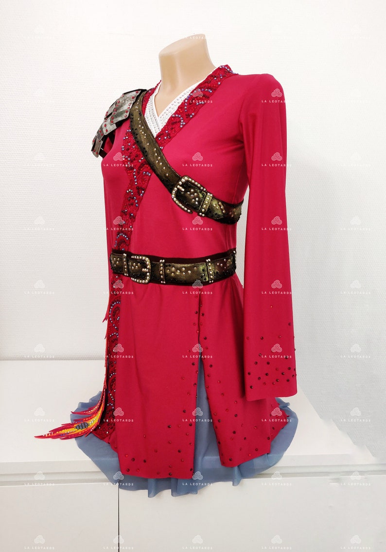 May include: A red figure skating dress with long sleeves, a white lace collar, and a brown belt with a buckle. The dress is decorated with rhinestones and has a gold and black trim. The dress has a short skirt with a blue tulle underskirt.