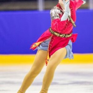 May include: A young woman figure skater wearing a red, blue, and gold costume with a red headband. She is performing a jump on the ice.
