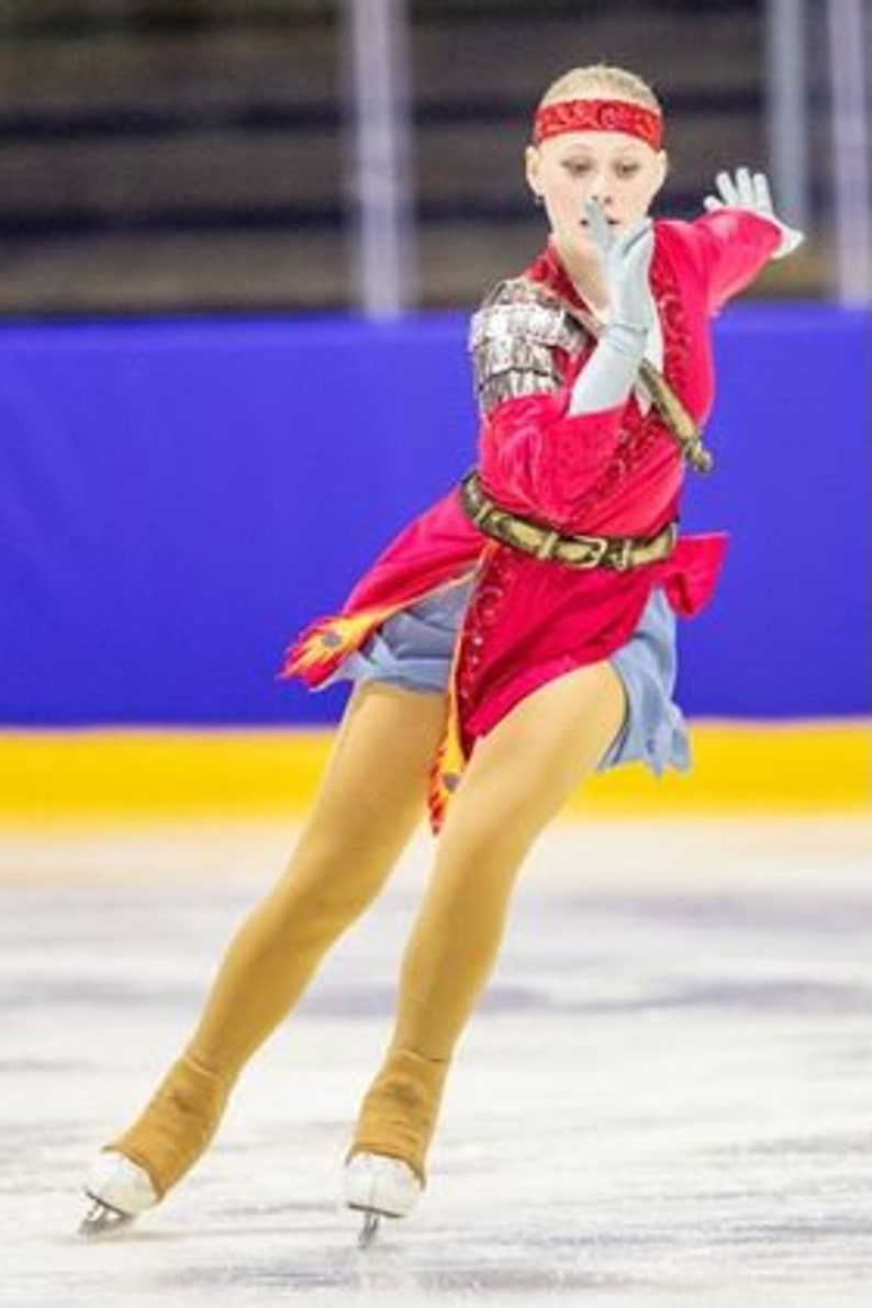 May include: A young woman figure skater wearing a red, blue, and gold costume with a red headband. She is performing a jump on the ice.