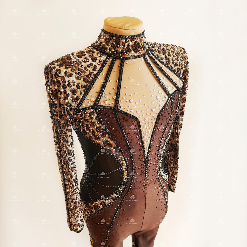 May include: A brown and black long-sleeved leotard with a leopard print design. The leotard has a high neckline and a mesh panel in the front that is decorated with rhinestones.