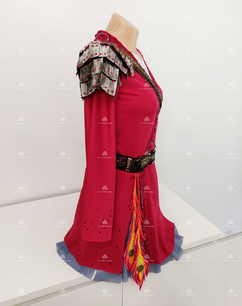 May include: A red figure skating dress with long sleeves, a silver shoulder piece, a brown belt, and a colorful feather design on the skirt.