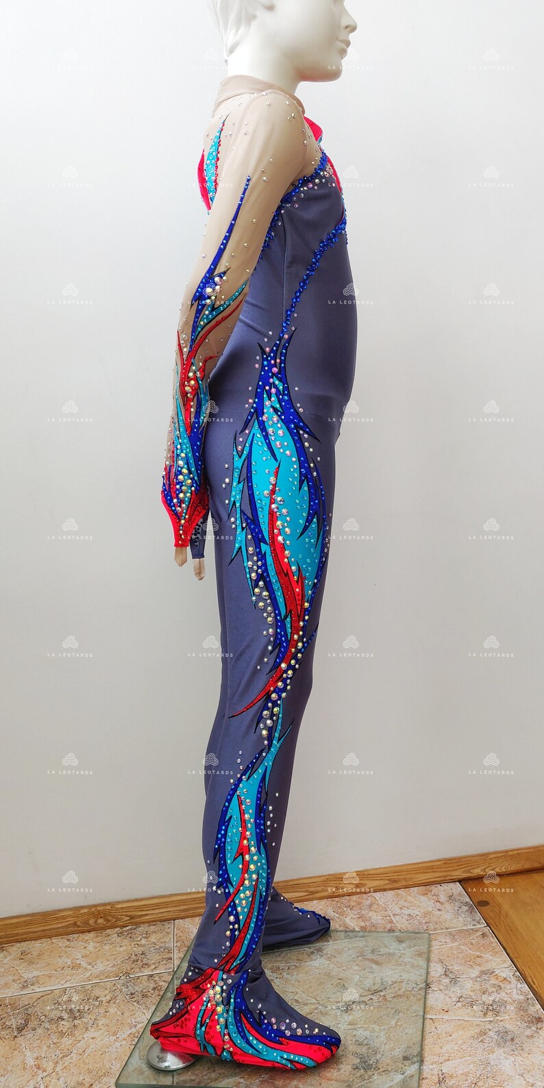 Op de afbeelding: Een grijze maillot met lange mouwen en een kleurrijk ontwerp met blauwe, rode en turquoise accenten. De maillot is versierd met strass en heeft een aansluitende pasvorm.