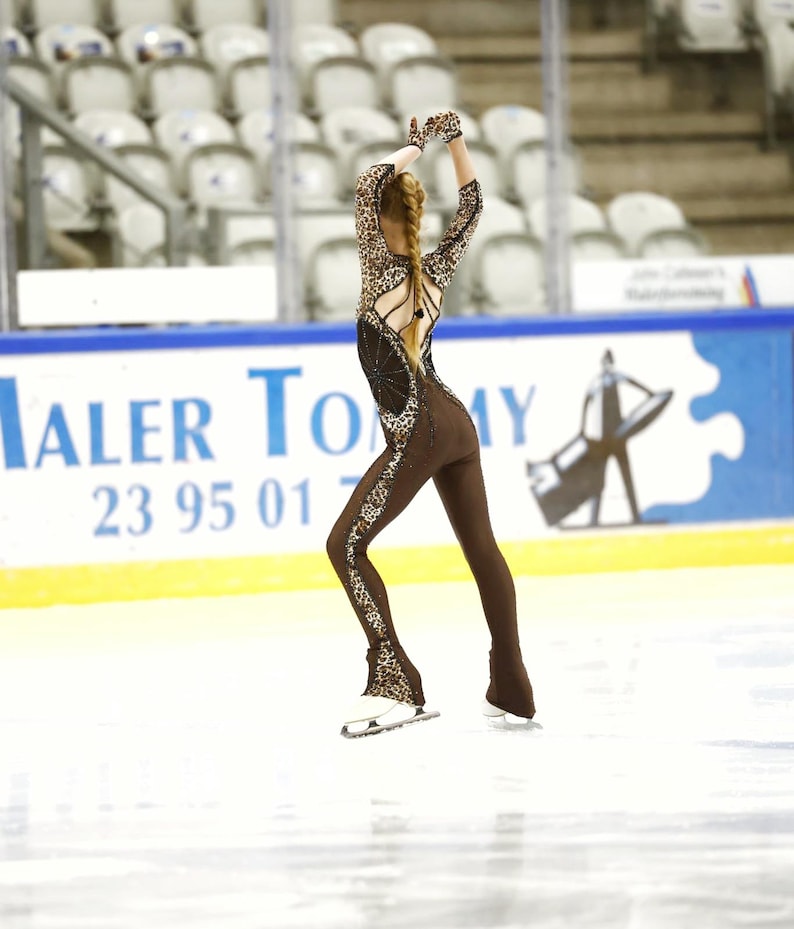 May include: A figure skater wearing a brown and black animal print leotard with a long braid. The skater is on the ice with one leg extended and the other leg bent. The skater is wearing black gloves and white skates.