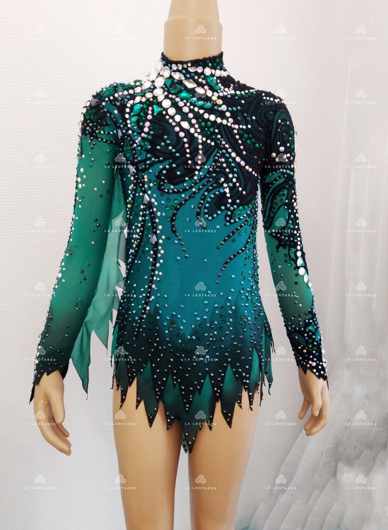 May include: A teal and black leotard with long sleeves, adorned with rhinestones. The leotard has a high neckline and a decorative pattern on the bodice and sleeves. The bottom hem has a jagged, leaf-like design. The brand "LA Leotards" is visible.