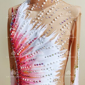 K&ouml;nnte beinhalten: Ein wei&szlig;-rosa Eiskunstlauf-Leotard mit Federdesign und Perlenbesatz. Der Leotard hat lange &Auml;rmel und einen hohen Kragen.