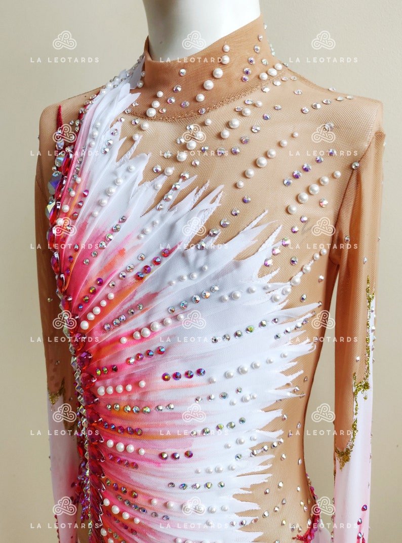K&ouml;nnte beinhalten: Ein wei&szlig;-rosa Eiskunstlauf-Leotard mit Federdesign und Perlenbesatz. Der Leotard hat lange &Auml;rmel und einen hohen Kragen.