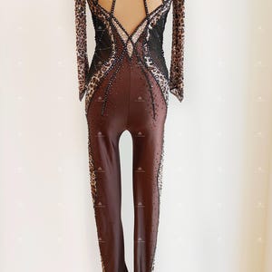 May include: A brown and black long-sleeved leotard with a leopard print design. The leotard has a low back with a mesh panel and a cutout design. The leotard is embellished with rhinestones.