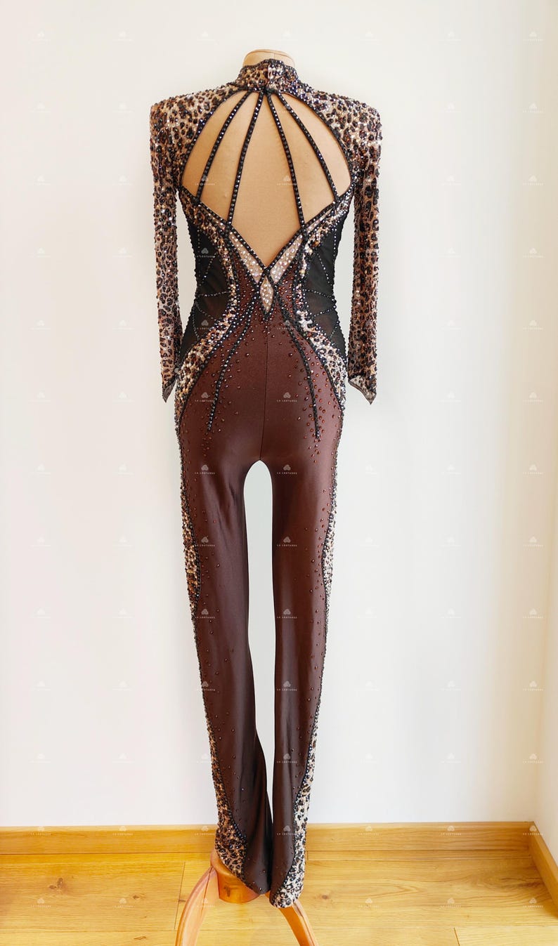 May include: A brown and black long-sleeved leotard with a leopard print design. The leotard has a low back with a mesh panel and a cutout design. The leotard is embellished with rhinestones.