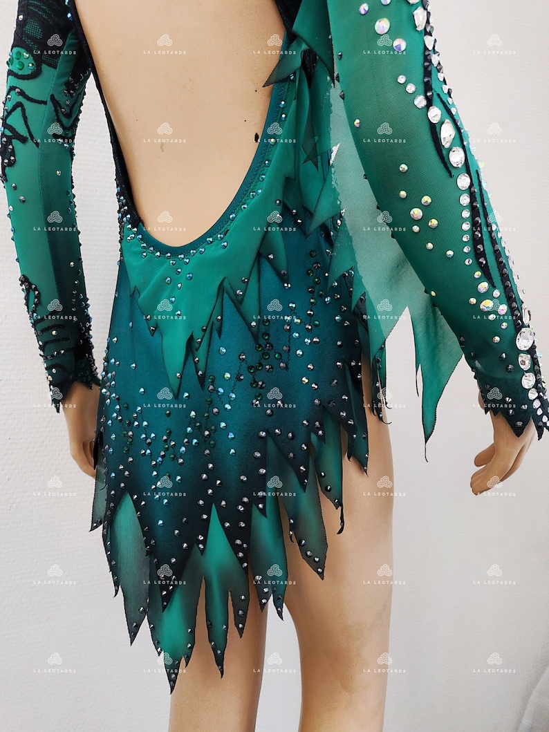 May include: A green and black leotard with long sleeves, adorned with rhinestones. The backless design showcases a gradient of green shades and a jagged, leaf-like hem. The sleeves are sheer, and the garment is designed for performance.